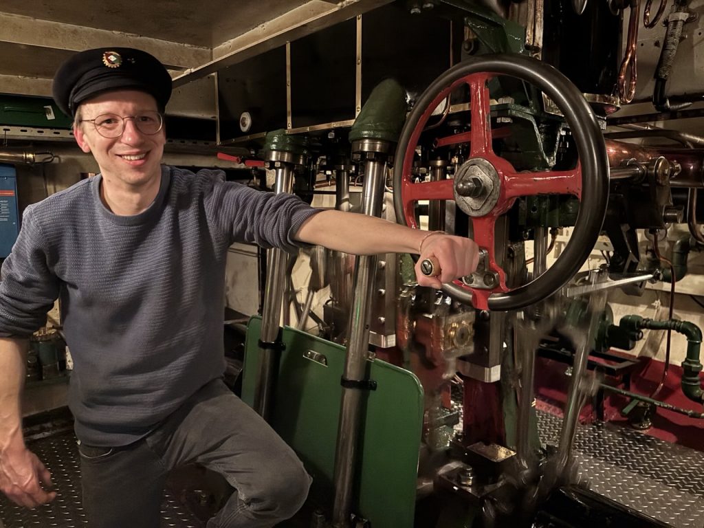 Mann mit Brille lächelt in Kamera, Hand an rotem Ventilrad im Maschinenraum eines historischen Dampfschiffs
